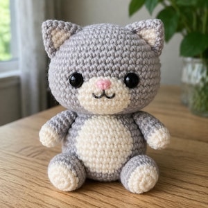 May include: A handmade crochet cat doll in gray and cream colors. The cat has large black eyes, a pink nose, and a smiling mouth. The doll is sitting on a wooden surface. The cat's ears and paws are cream-colored.