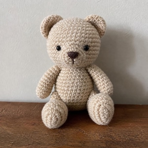 May include: A handmade, beige crochet teddy bear with a brown nose and black eyes. The bear is sitting upright with its arms and legs extended. The bear is made of a soft, textured yarn.