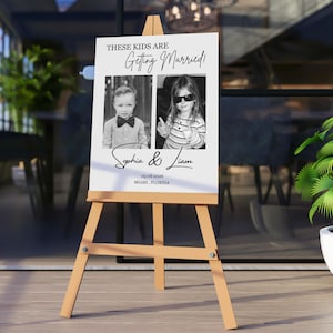 Puede incluir: Un caballete de madera exhibe un letrero blanco con el texto "THESE KIDS ARE Getting Married!" en escritura negra. Se presentan dos fotos en blanco y negro de ni&ntilde;os. El letrero incluye los nombres "Sophia & Liam" y la fecha y el lugar.
