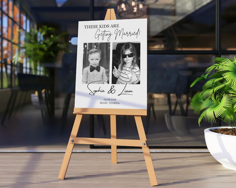 Puede incluir: Un caballete de madera exhibe un letrero blanco con el texto "THESE KIDS ARE Getting Married!" en escritura negra. Se presentan dos fotos en blanco y negro de ni&ntilde;os. El letrero incluye los nombres "Sophia & Liam" y la fecha y el lugar.