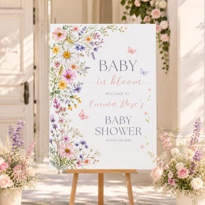 May include: A white rectangular sign on a wooden easel featuring a watercolor floral arrangement of pink, yellow, and purple wildflowers with butterflies. The text reads: BABY in bloom, WELCOME TO Emma Rose's BABY SHOWER, AUGUST 20, 20XX.