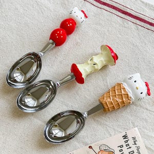 May include: Three stainless steel ice cream scoops with decorative handles. One handle features a white cat head with a red bow, another has red spheres, and the third is an apple core. The scoops are on a white surface.