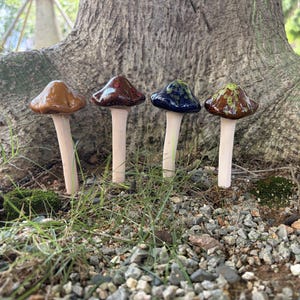 May include: Four ceramic mushroom sculptures with smooth, light-colored stems and glossy, rounded caps in solid brown, dark reddish-brown, deep blue with green speckles, and brown with green and yellow mottled patterns, arranged in a row.