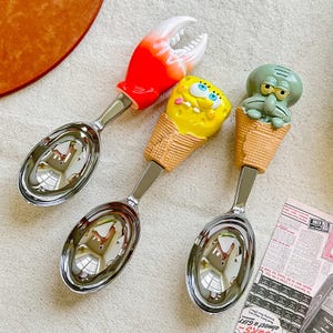 May include: Three ice cream scoops with character handles. One has a red and white crab claw, another a yellow SpongeBob in an ice cream cone, and the third a green Squidward in a cone. The scoops are made of stainless steel.