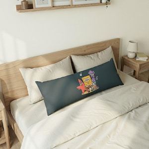 May include: A long, rectangular pillow with a dark teal cover featuring a cartoon graphic of SpongeBob SquarePants, Patrick Star, and Squidward Tentacles. The pillow is placed on a bed with cream-colored bedding.