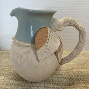 May include: A handmade ceramic pitcher with a light blue glaze on the top and a textured, off-white body. The pitcher has a leaf design on the side and a sculpted handle.