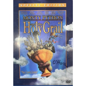 Może przedstawiać: Okładka DVD do filmu Monty Python i Święty Graal. Okładka przedstawia niebieskie tło z dużym obrazem złotego kielicha trzymanego przez rękę. Tytuł filmu jest napisany białymi literami na czarnym tle. Nazwiska aktorów są wymienione pod tytułem.