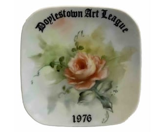 Op de afbeelding: Vierkante keramische plaat met een handgeschilderde perzikroos en groene bladeren. De plaat heeft de tekst "Doylestown Art League" in zwart schrift bovenaan en "1976" onderaan. De achtergrond is een zacht, gemengd aquareleffect.