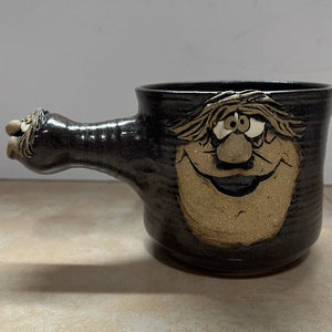 Peut inclure: Une tasse en céramique brune avec un visage fantaisiste et une longue poignée. Le visage a une barbe brune, un grand nez et un large sourire. La tasse est émaillée d'une couleur brun foncé avec une finition mouchetée.