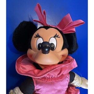 Puede incluir: Mu&ntilde;eca de peluche de Minnie Mouse con vestido, cuello y lazo de sat&eacute;n rosa. La mu&ntilde;eca tiene orejas negras, nariz negra y cara bronceada. Los brazos de la mu&ntilde;eca son blancos con ribetes dorados.