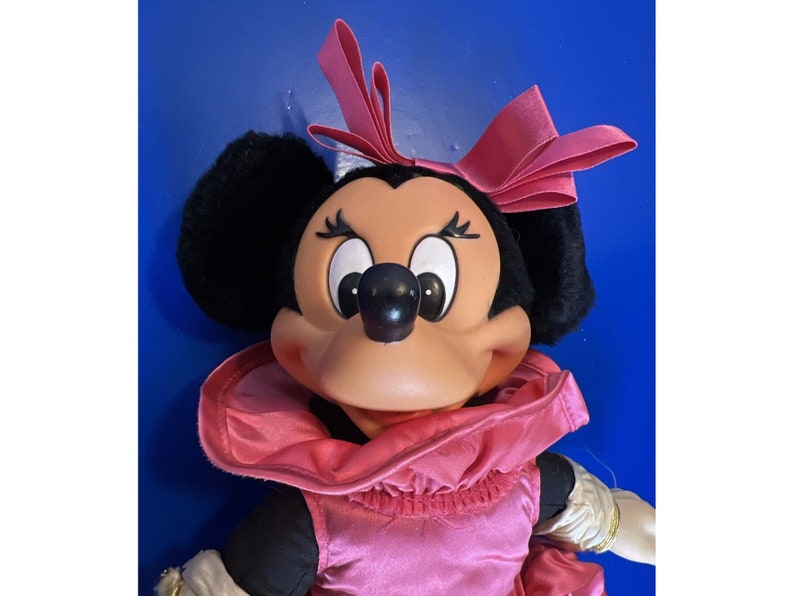 Puede incluir: Mu&ntilde;eca de peluche de Minnie Mouse con vestido, cuello y lazo de sat&eacute;n rosa. La mu&ntilde;eca tiene orejas negras, nariz negra y cara bronceada. Los brazos de la mu&ntilde;eca son blancos con ribetes dorados.