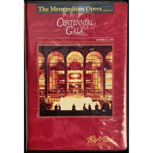 Pode incluir: Uma capa de DVD vermelha para a Gala do Centenário da Ópera Metropolitana, 22 de outubro de 1983. A capa apresenta o interior de um edifício com janelas arqueadas e uma fonte. O texto "Centennial Gala" é exibido em destaque.