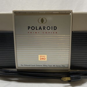 May include: A vintage Polaroid Print Copier, model 240, in beige and black with a silver front panel. The front panel has the Polaroid logo and the text "Print Copier". The copier is designed for use with Polaroid Land Cameras using Type 40 series film.