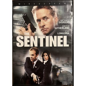 Puede incluir: Caja de DVD de la película "The Sentinel" con Michael Douglas, Kiefer Sutherland y Eva Longoria. La portada muestra a Michael Douglas sosteniendo un arma, con el título en letras blancas grandes.