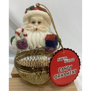 May include: A gold wire mesh candy ornament with a Santa Claus figurine on top. The ornament has a red tag that says "Ameri-Suisse Candy Ornament".