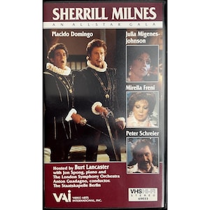 Pode incluir: Fita VHS para "Sherrill Milnes: An All-Star Gala" com Placido Domingo. A capa tem imagens de artistas e texto. A fita está em uma caixa preta com uma borda marrom e o logotipo VHS-Hi-Fi Stereo.