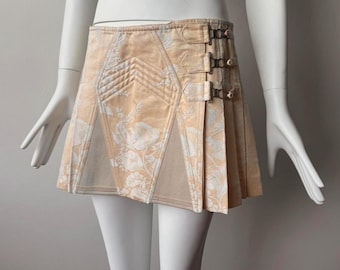 May include: A beige mini skirt with a floral pattern and pleats. The skirt features decorative stitching, a buckle closure, and a contrasting panel. The skirt is displayed on a mannequin.