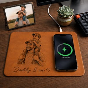 May include: A brown leather wireless charging pad with a custom engraved image of a father and child. A smartphone is charging on the pad, displaying a 67% charge. A framed photo and a succulent plant are also on the desk.