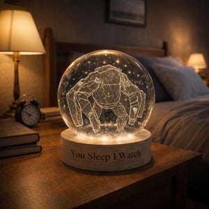 May include: A glowing glass sphere night light with a detailed etched design of a creature. The sphere sits on a wooden base with the text "You Sleep I Watch". The light emits a warm glow, perfect for a bedroom setting.