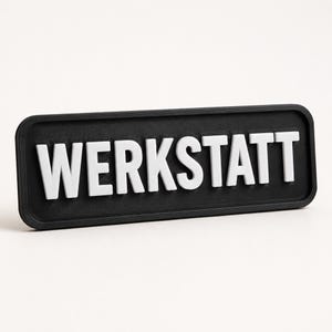 May include: A rectangular black sign with a raised border featuring the white embossed text WERKSTATT in a bold sans-serif font. The surface of the black base has a fine, linear textured finish, while the white letters are smooth and matte.