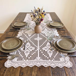 May include: A wooden dining table set with a cream-colored crocheted table runner, four place settings featuring speckled olive-green ceramic plates, silver cutlery, and clear glasses. A central ceramic vase holds a bouquet of dried flowers.