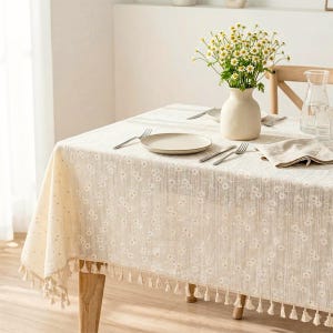 Puede incluir: Un mantel beige con un patrón de margaritas blancas y flecos en los bordes. El mantel cubre una mesa decorada con un jarrón de cerámica blanca que contiene un ramo de margaritas, dos platos blancos, dos tenedores y una servilleta doblada.