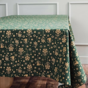 Puede incluir: Un mantel de color verde oscuro con un patrón repetitivo de ilustraciones de líneas en color dorado. El estampado incluye varios adornos colgantes, campanas estilizadas, copos de nieve y pequeños pinos distribuidos por la tela.