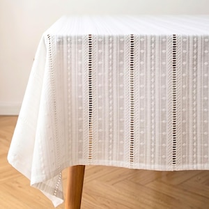 May include: A white tablecloth with a detailed, textured pattern. The design features vertical stripes with dotted and geometric accents. The cloth is draped over a wooden table, showcasing its elegant, decorative style.