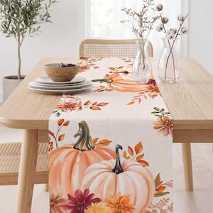 Pode incluir: Um caminho de mesa retangular com um padrão estampado de abóboras laranja e brancas, folhas de outono e várias flores em tons de amarelo, laranja e vermelho escuro sobre um fundo de tecido bege claro, exibido numa mesa de jantar de madeira.