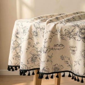 Könnte beinhalten: Cremefarbene Blumendecke mit schwarzem Blumenmuster. Die Tischdecke hat einen schwarzen Quastenbesatz am Rand. Das florale Design umfasst große Blumen und zarte Blätter und schafft eine elegante Ästhetik.
