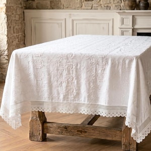 Puede incluir: Un mantel rectangular blanco con un intrincado diseño floral y un delicado borde de encaje. El mantel está drapeado sobre una mesa de madera rústica, creando un aspecto clásico y elegante. La tela parece ser lino o un material similar.