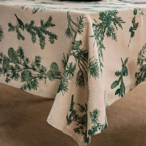 Puede incluir: Un mantel de color beige con un patrón repetitivo de ramas de pino, agujas y piñas en color verde oscuro. La tela tiene una textura tejida visible y las ilustraciones botánicas están impresas en una tinta de color verde bosque.