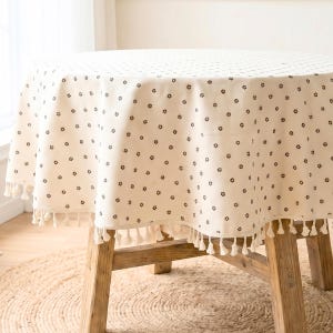 Peut inclure: Une nappe ronde crème avec un motif répété de petites formes florales brun foncé. Le bord est orné d'une frange de pompons blancs. La nappe est drapée sur une table en bois.