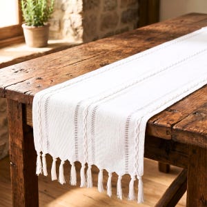 Puede incluir: Camino de mesa blanco con tejido texturizado y diseño de rayas verticales. El camino de mesa tiene un fleco de borlas a lo largo del borde inferior. El camino de mesa se exhibe sobre una mesa de madera rústica.