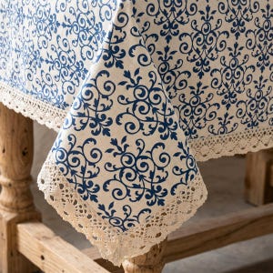 May include: A cream tablecloth featuring a repeating blue floral design and a delicate lace trim. The cloth is draped over a wooden table, highlighting its pattern and texture. The design includes swirling vines and leaves.