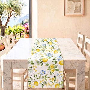 Op de afbeelding: Een witte tafelloper met een herhalend aquarelpatroon van gele citroenen, groene bladeren en kleine blauwe en gele bloemen. De loper ligt in het midden op een witte marmeren tafel en toont een botanisch ontwerp met levendige fruit- en bloemmotieven.