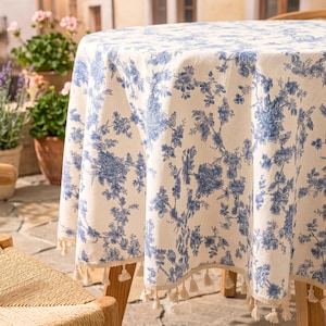 Op de afbeelding: Een ronde tafelkleed met een crèmekleurige achtergrond en een blauw bloemmotief. De tafelkleed heeft een franje van kleine kwastjes langs de rand. Het ligt op een houten tafel.