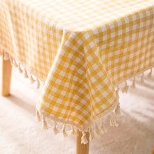 May include: A yellow and white checkered tablecloth with a fringe of tassels along the edge. The tablecloth is draped over a wooden table, creating a cozy and inviting atmosphere. The fabric has a classic gingham pattern.