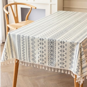 Pode incluir: Uma toalha de mesa retangular com base na cor creme e listras verticais de padrões geométricos pretos, incluindo diamantes, triângulos e pontos. As bordas do tecido são finalizadas com uma fileira decorativa de pequenas borlas em tom off-white.