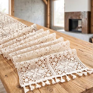 Op de afbeelding: Crèmekleurige kanten tafelloper met een gedetailleerd openwerk ontwerp. De loper is over een houten tafel gedrapeerd en toont zijn ingewikkelde patronen en delicate kwastjes langs de randen. Het voegt elegantie toe.