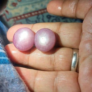 Könnte beinhalten: Zwei runde, im Cabochon-Schliff geschliffene rosa Sternsaphire, die jeweils einen deutlichen sechsstrahligen Sterneffekt auf ihrer Oberfläche zeigen. Die Edelsteine sind glatt, poliert und weisen einen sanften, durchscheinenden Rosaton auf.