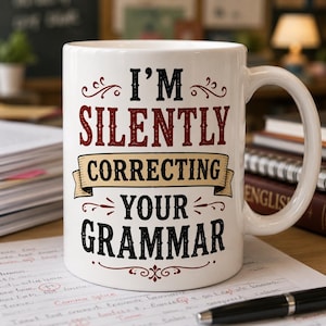 Puede incluir: Taza de cerámica blanca con el texto "I'M SILENTLY CORRECTING YOUR GRAMMAR" en letras negras y granates. La taza tiene un asa curva y está sobre una superficie de madera con papeles y un bolígrafo.