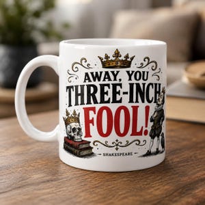 Puede incluir: Una taza de cerámica blanca con un gráfico de corona dorada sobre el texto negro "AWAY, YOU THREE-INCH FOOL!" con la palabra "FOOL!" en rojo. Incluye una calavera con corona sobre dos libros y una figura histórica junto a un cuervo.