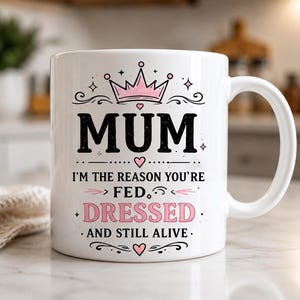 Pode incluir: Caneca de cerâmica branca com a palavra "MUM" em preto, uma coroa rosa e o texto "I'M THE REASON YOU'RE FED, DRESSED, AND STILL ALIVE." O design inclui corações e estrelas.