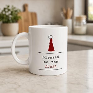 Op de afbeelding: Een witte keramische mok met een minimalistische rode illustratie van een figuur met een kapje en een lange jurk, met de zwarte en rode tekst "blessed be the fruit" gecentreerd tussen twee horizontale zwarte lijnen op de voorzijde.
