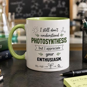 May include: White ceramic mug with a green handle and interior. The mug features the text "I still don't understand PHOTOSYNTHESIS but I appreciate your ENTHUSIASM." with illustrations of a sun, leaves, and a chemical formula. A black pen and sticky notes are in the foreground.