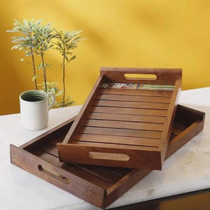 Könnte beinhalten: Zwei rechteckige Serviertabletts aus Holz mit Lattenboden und Griffaussparungen. Jedes Tablett verfügt über eine dekorative Keramikflieseneinlage mit bunten Mustern an einer Kante. Die Tabletts sind gestapelt und eine weiße Tasse steht daneben.