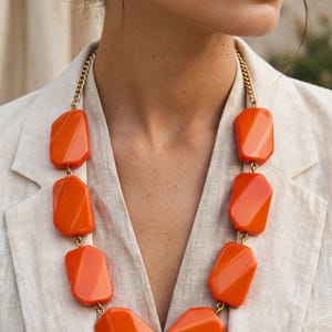 Puede incluir: Un collar llamativo con grandes cuentas de resina facetadas de color naranja brillante conectadas por una cadena de metal dorado. Las cuentas están dispuestas en un patrón simétrico, creando un aspecto audaz sobre la tela texturizada clara.