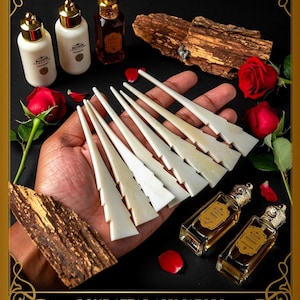May include: A collection of bone attar applicators, alongside perfume bottles, wood, and red roses. The applicators are white and feature a distinctive, stepped design. The bottles have gold detailing. The image is set against a dark backdrop.