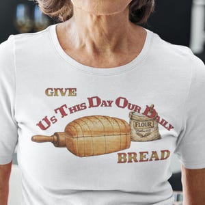 May include: White t-shirt featuring a graphic with the text "Give Us This Day Our Daily Bread." The design includes a loaf of bread, a rolling pin, and a bag of flour. The text and graphics are in shades of brown and gold.